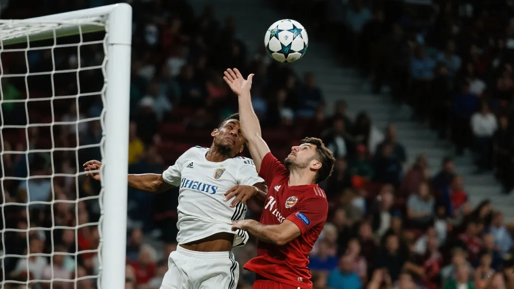 禁区内球员起跳争顶，皮球贴近手臂，观众席模糊成光斑的紧张画面